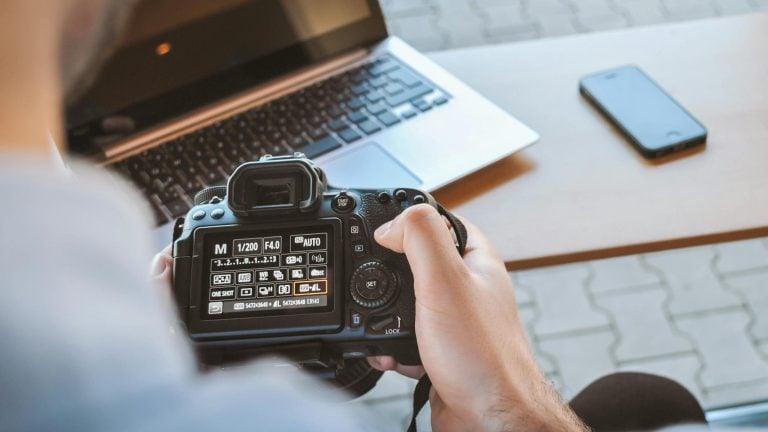 Close-up of hands holding DSLR camera at a workspace with laptop and smartphone.